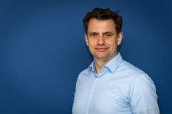 Eine Person mit kurz geschnittenem, dunklem Haar trägt ein hellblaues, langärmliges Hemd mit Kragen. Der Oberkörper ist leicht seitlich gedreht. Der Hintergrund ist einfarbig dunkelblau. Das Gesicht ist unkenntlich gemacht.